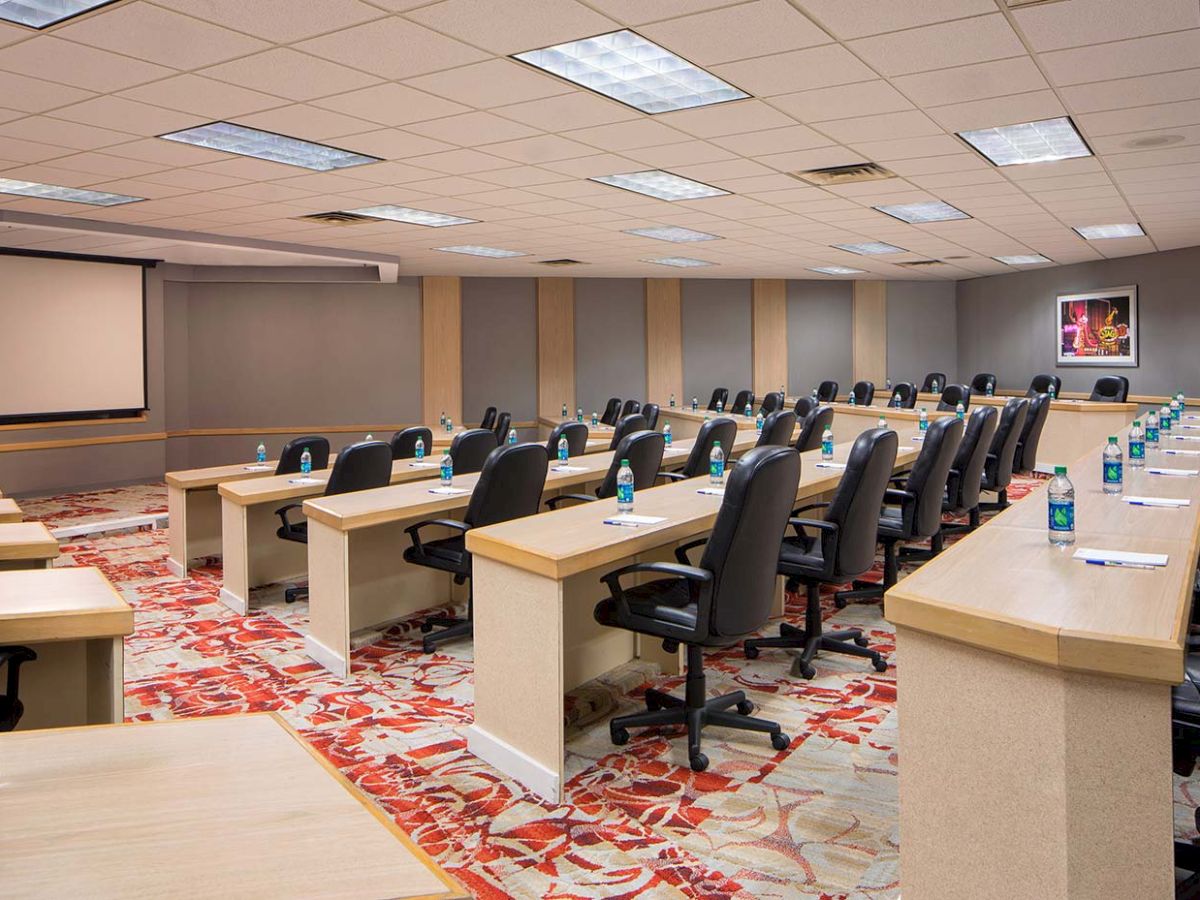 A conference room with rows of tables and chairs, bottles of water, notepads, and a projection screen at the front, under bright lighting.