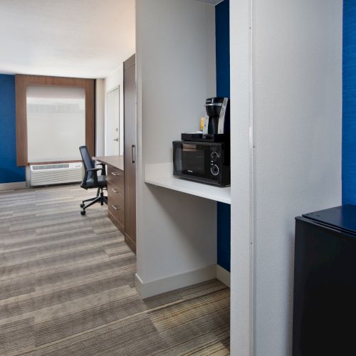 A hotel room with a microwave, coffee maker, mini-fridge, desk, chair, and window with blue accent walls and modern decor.