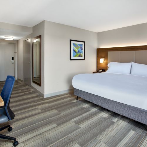 A hotel room with a large bed, desk, chair, wall art, TV, and mirror, featuring neutral tones and modern decor.