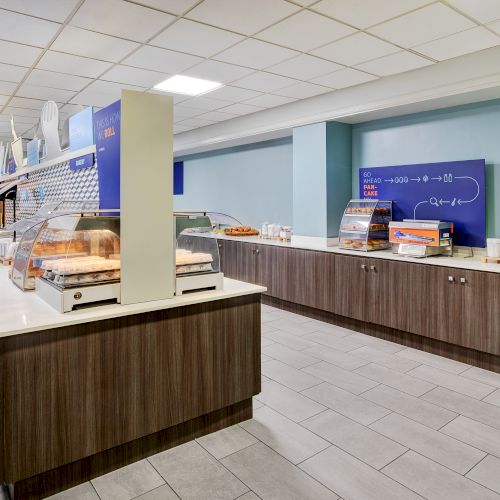 The image shows a bright cafeteria with food display cases and counters set up for self-service in a clean, modern setting.