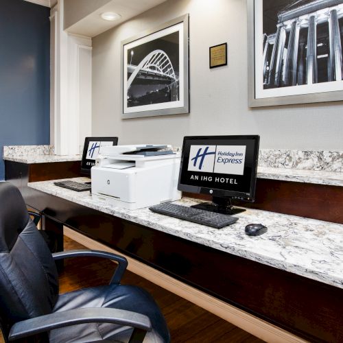A business center with two computers, a printer, and two chairs on a marble countertop beneath framed black-and-white photographs.