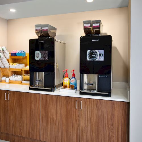 The image shows a countertop with two coffee machines, cups, lids, and condiments alongside a waste bin sign on the wall.