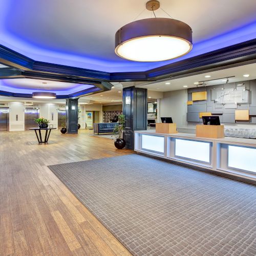 The image shows a modern hotel lobby with a reception desk, stylish lighting, wood flooring, and a spacious seating area.