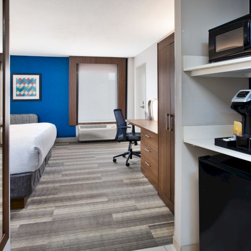 This image shows a modern hotel room with a bed, desk, chairs, microwave, and coffee maker, featuring a blue accent wall and wood flooring.