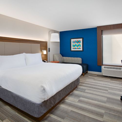 A modern hotel room with a king-size bed, desk, chair, couch, blue accent wall, and minimalistic decor.