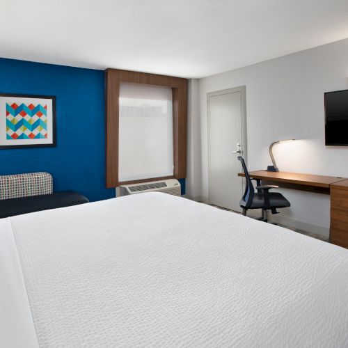 This image shows a modern hotel room with a bed, a desk with a chair, a flat-screen TV, and a colorful wall painting.