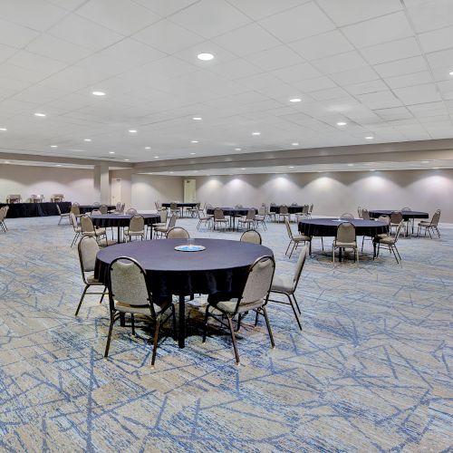 The image shows a large, empty conference room with round tables and chairs, decorated in neutral tones with patterned carpeting.