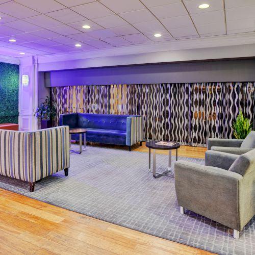 A modern lounge area with sofas, armchairs, tables, and decorative wall art, featuring warm lighting and a patterned carpet on the floor.