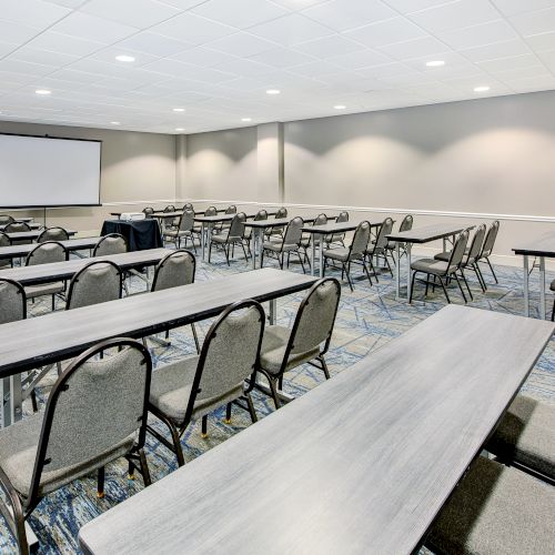 A conference room with several rows of tables and chairs arranged facing a projector screen on the wall.