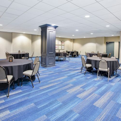 A conference room with round tables covered in black cloths, surrounded by chairs, on a blue carpet, and with neutral-colored walls.