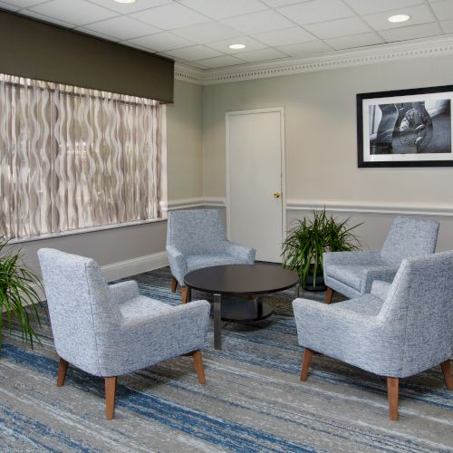 A modern waiting room with four armchairs around a small table, plants, a window with curtains, and a framed picture on the wall.