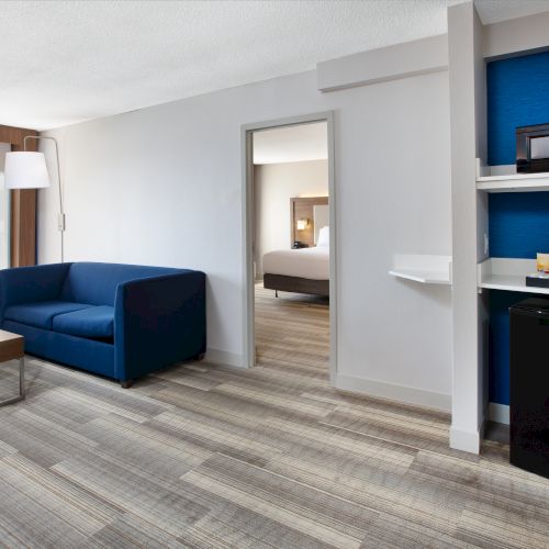 The image shows a modern hotel room with a blue sofa, a coffee table, microwave, mini-fridge, and sleek wooden flooring.