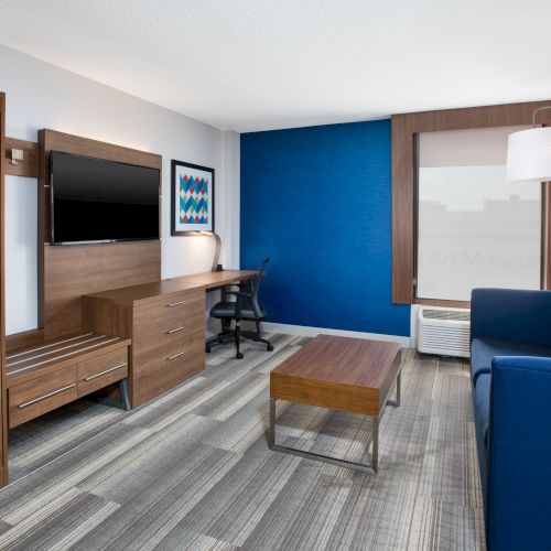 A modern hotel room with a TV, desk, and couch. The decor features blue and wood tones, creating a clean, contemporary atmosphere.