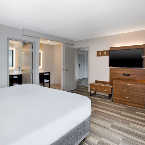 The image shows a modern hotel room with a bed, wall art, desk, chair, TV, dresser, and an open door leading to a bathroom area.
