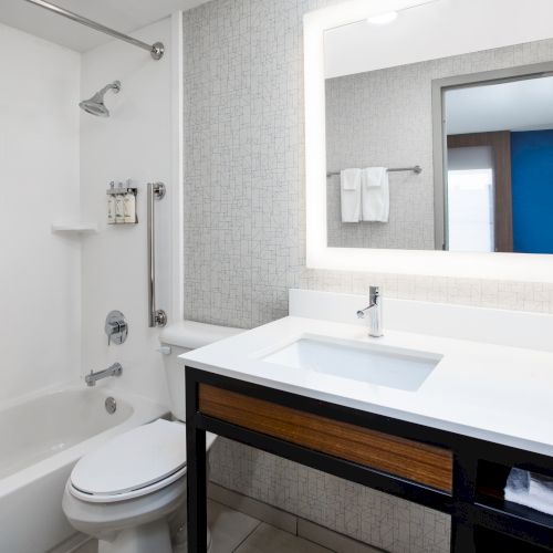 A modern bathroom with a bathtub, toilet, and vanity. It features a large illuminated mirror and minimalistic design elements.
