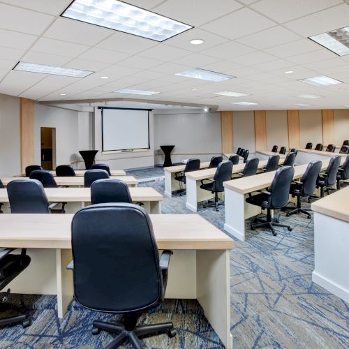 The image shows a modern conference room with rows of desks and chairs facing a projection screen at the front of the room.