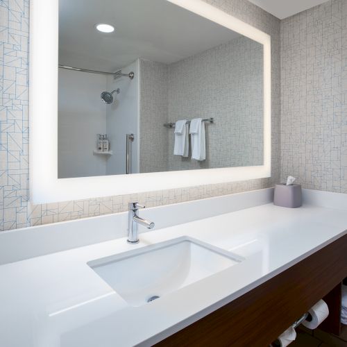 A modern bathroom features a large illuminated mirror, white countertop with a sink, faucet, and neatly arranged towels and toiletries.
