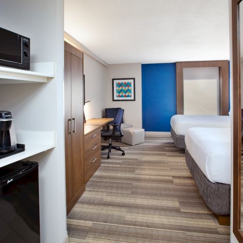 A modern hotel room with two beds, a desk, TV, and kitchenette area including a coffee maker and microwave.