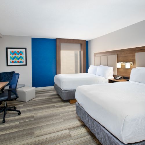 A modern hotel room with two beds, a desk with a chair, a TV, and a framed picture on the wall, featuring neutral and blue tones.