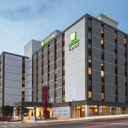 The image shows a modern multi-story hotel building at dusk, with illuminated windows and bustling streets.