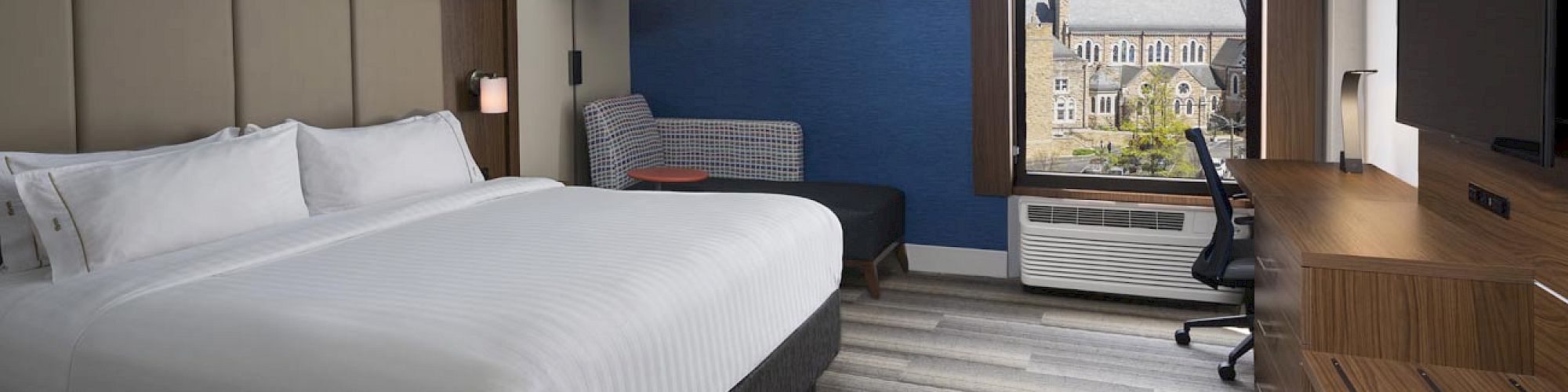 The image shows a modern hotel room with a bed, desk, chair, TV, and window with a cityscape view.