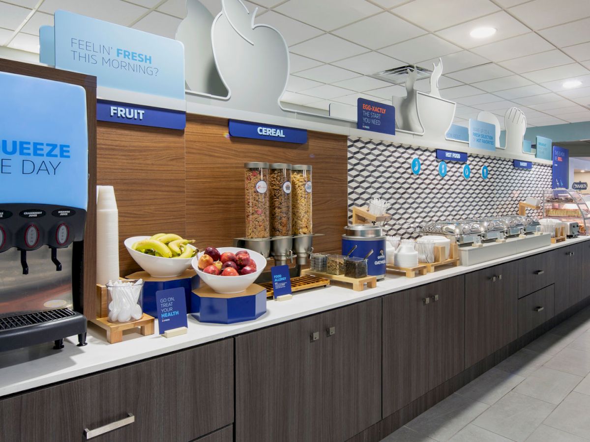 The image shows a breakfast buffet with juice dispensers, a fruit display, cereal containers, and various breakfast items on a countertop.