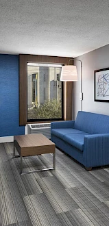 The image shows a modern hotel room with a blue sofa, desk, and a view into a bedroom area with a bed and window.