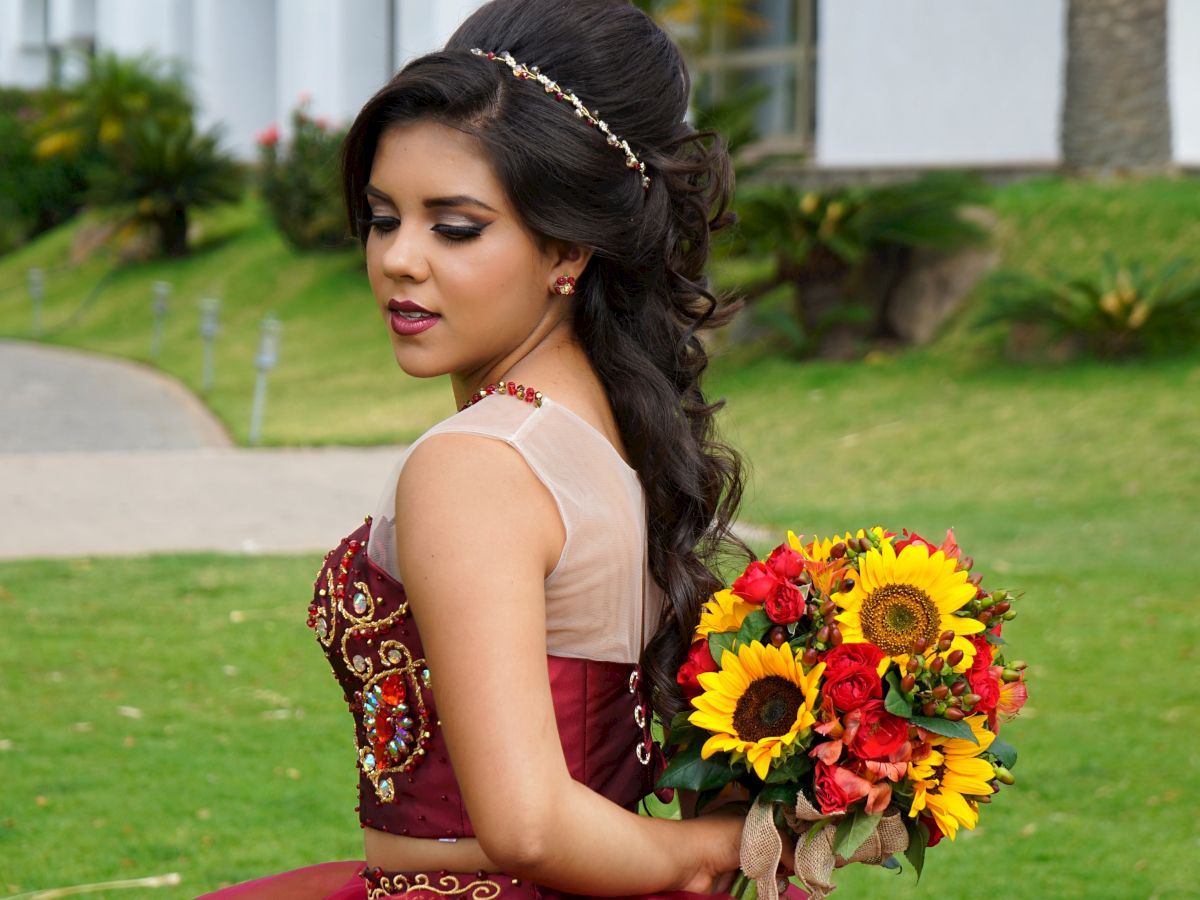 A young woman in a red gown holds a bouquet of sunflowers and red flowers, standing on grass near a white building.