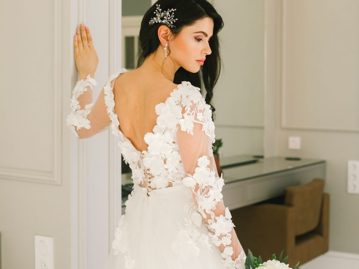 A woman in a white floral wedding dress holds a bouquet, standing near a wall, with a decorative hairpiece in an elegant room.