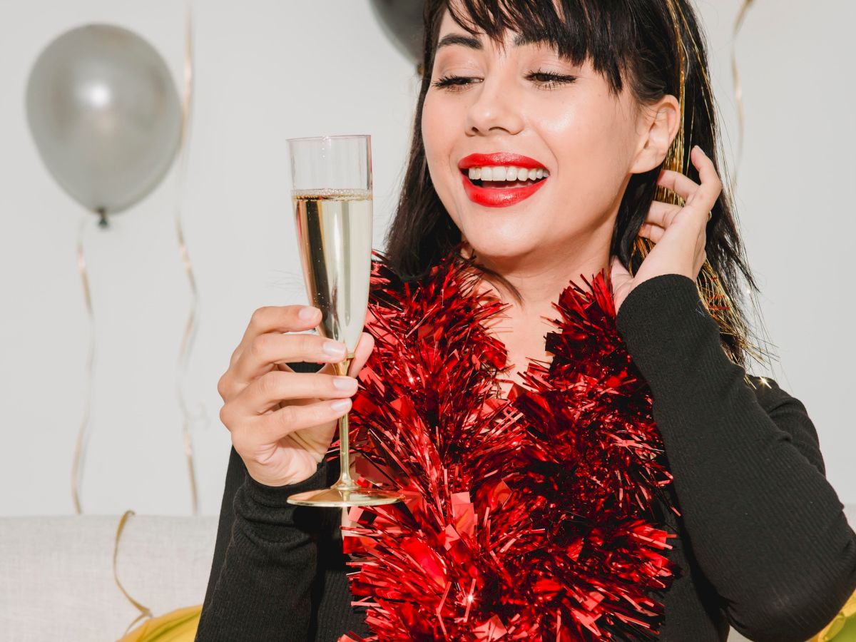 A person is joyfully holding a glass, wearing a red tinsel scarf. Balloons are in the background.