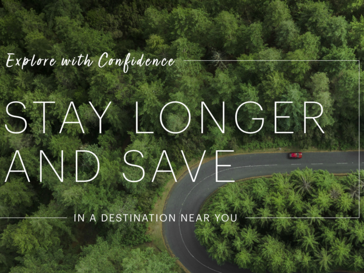 Aerial view of a car on a winding road lined with trees, featuring text: "Explore with Confidence. Stay Longer and Save. In a destination near you."