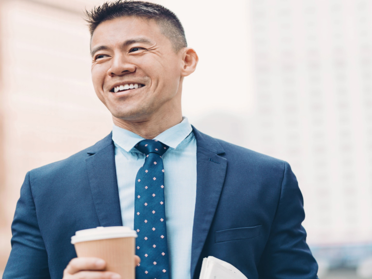 A person in a suit, smiling, holds a coffee cup and a newspaper outdoors, with buildings in the background.