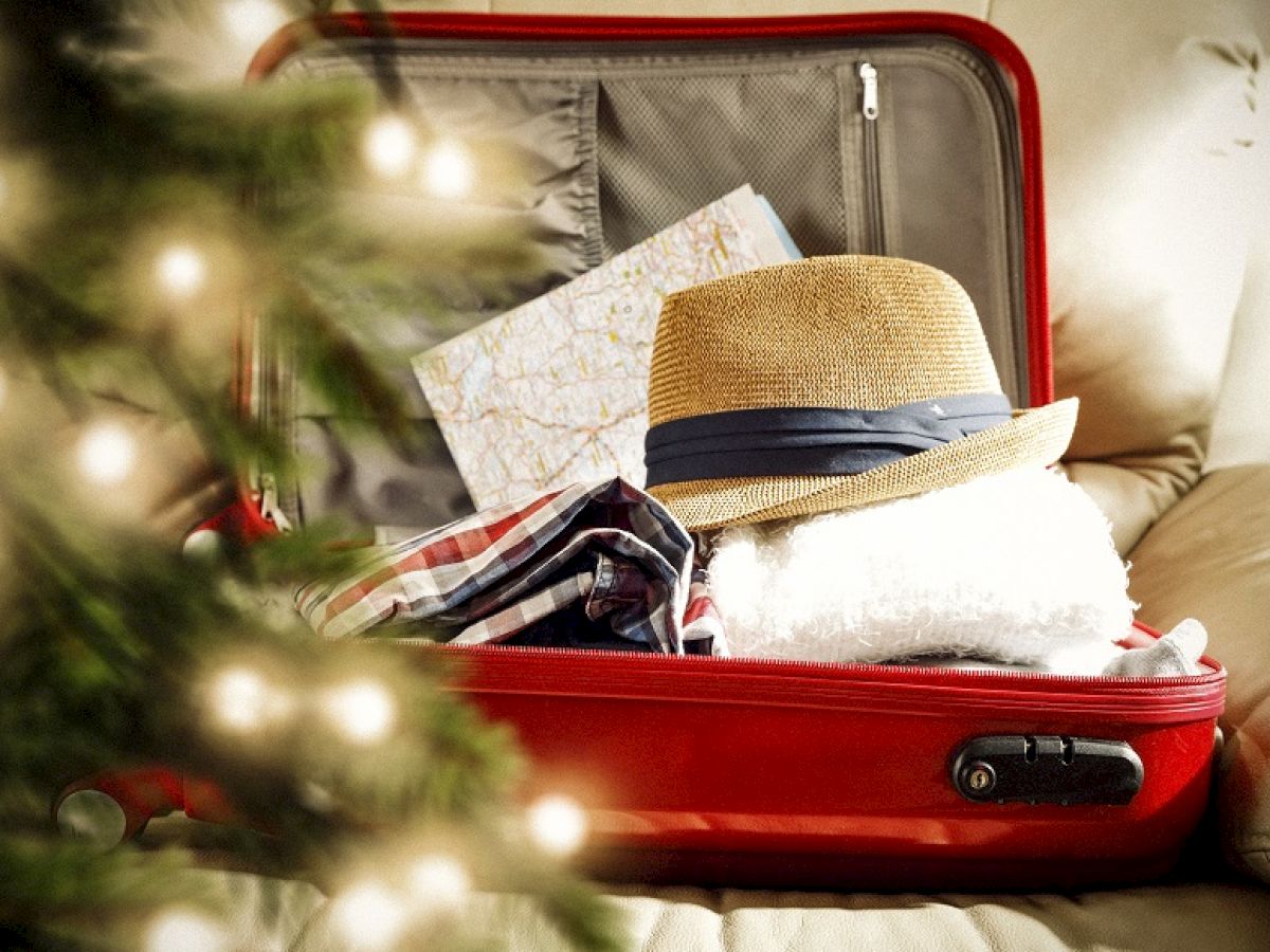 An open red suitcase with clothes, a straw hat, and a map is packed, next to a softly lit Christmas tree on a couch.