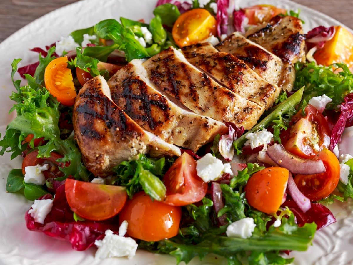 Grilled chicken breast is served on a bed of fresh salad with tomatoes, mixed greens, onions, and crumbled cheese.