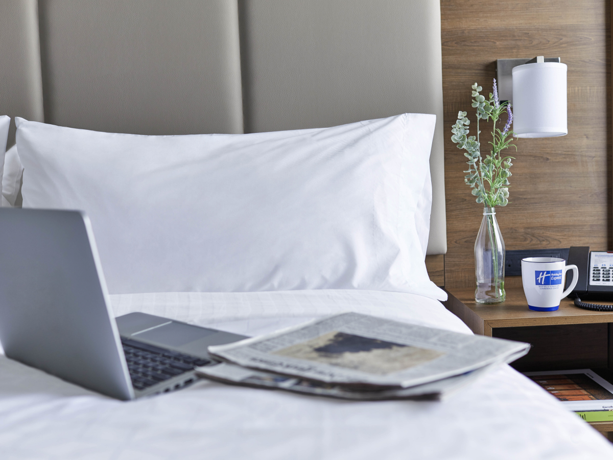 A bed with a laptop, newspapers, a cup, a phone, and a vase with flowers on the side table against a wooden and upholstered backdrop.