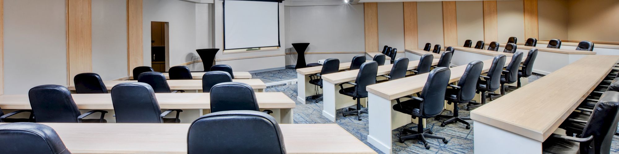 A modern conference room with several rows of desks and chairs facing a presentation screen, ready for a meeting or seminar.