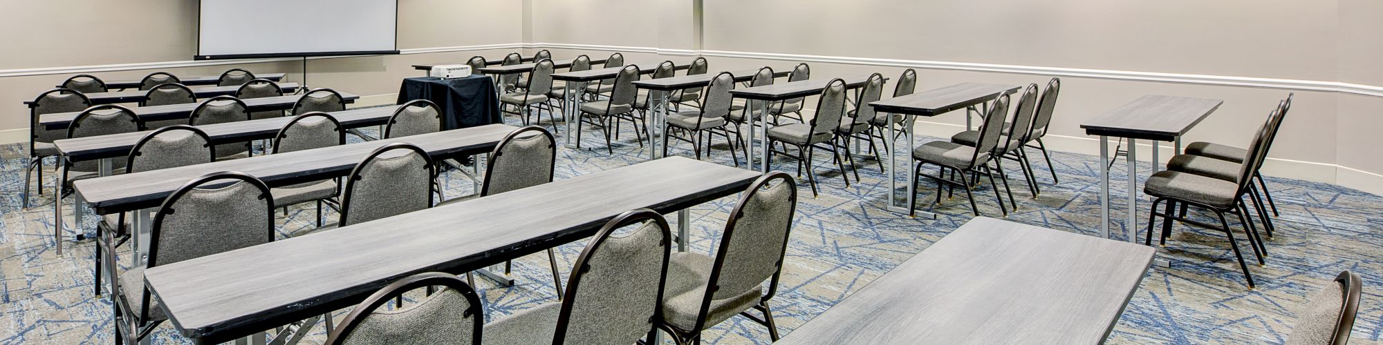 A conference room with grey tables and chairs is arranged in rows facing a screen, ready for a presentation or meeting.