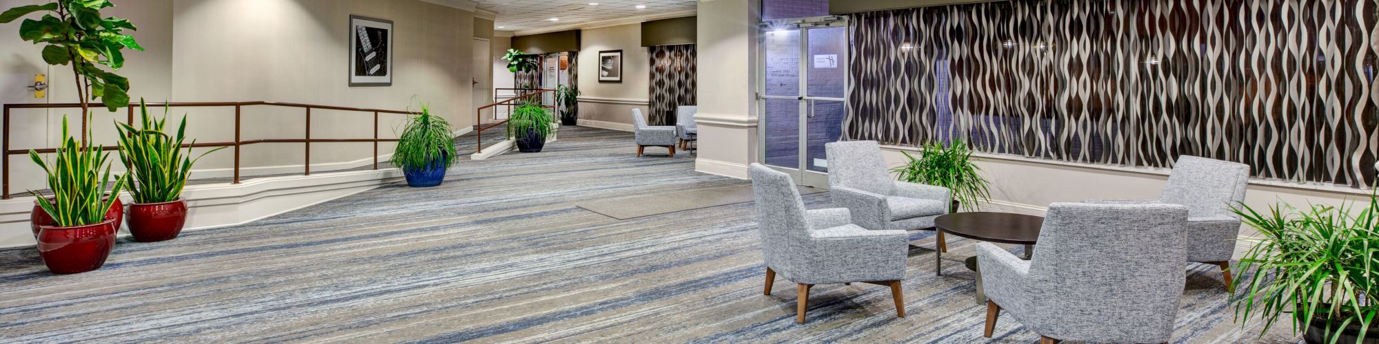 A modern lobby with a patterned carpet, seating area with chairs and tables, and potted plants for decoration, under ceiling lights.