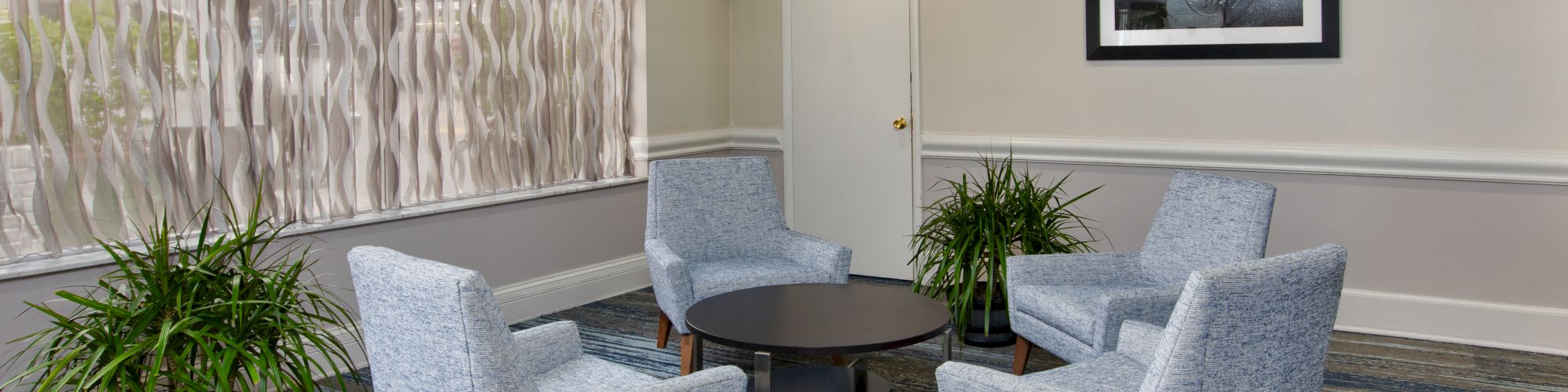 The image shows a seating area with four gray chairs around a round table, two potted plants, a window, and a photo on the wall.