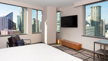 A modern hotel room with large windows showing a cityscape, featuring a bed, chair, TV, and a small table.
