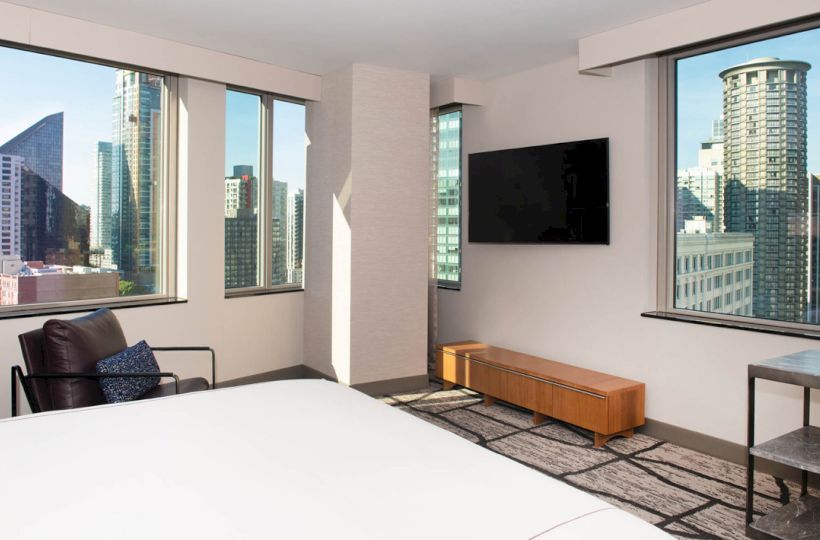 A modern hotel room with large windows showing a cityscape, featuring a bed, chair, TV, and a small table.