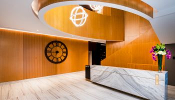 A modern lobby features a marble reception desk, large wall clock, wood paneling, and a winding staircase with globe-shaped lights.