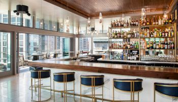 A modern bar with stylish seating, a variety of bottles on shelves, and large windows offering a city view.