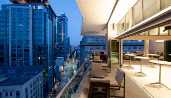 A modern balcony with seating overlooks a city street lined with high-rise buildings at dusk. The interior has tables and open spaces.