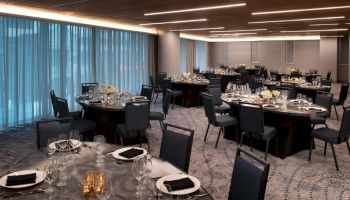 A well-lit banquet hall with round tables set for dining, featuring elegant glassware, black napkins, and flower centerpieces.