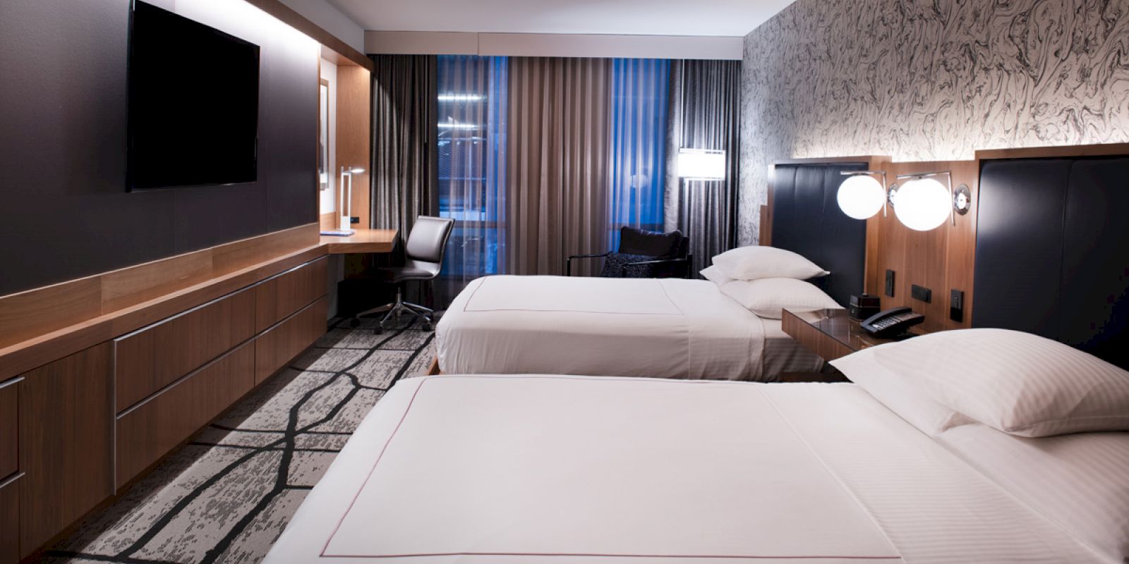 A modern hotel room with two beds, a wall-mounted TV, a desk, and decorative lighting, creating a cozy atmosphere.