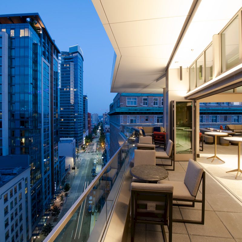 A modern cityscape with tall buildings, viewed from a stylish rooftop terrace with seating, under the evening sky.