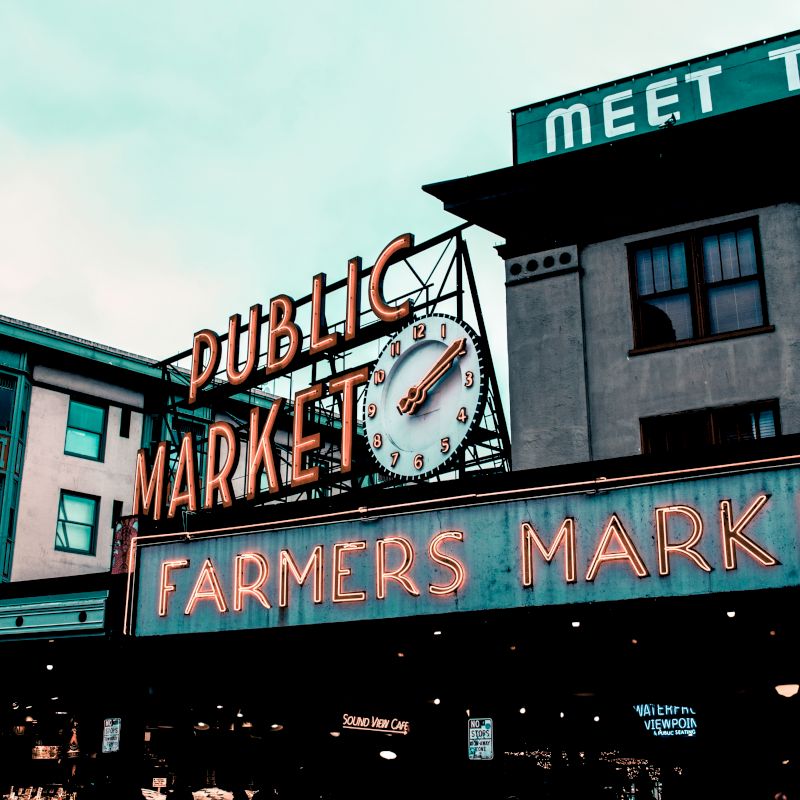 A neon sign reads "Public Market" and "Farmers Market," with a clock above, set against a multi-story building backdrop.