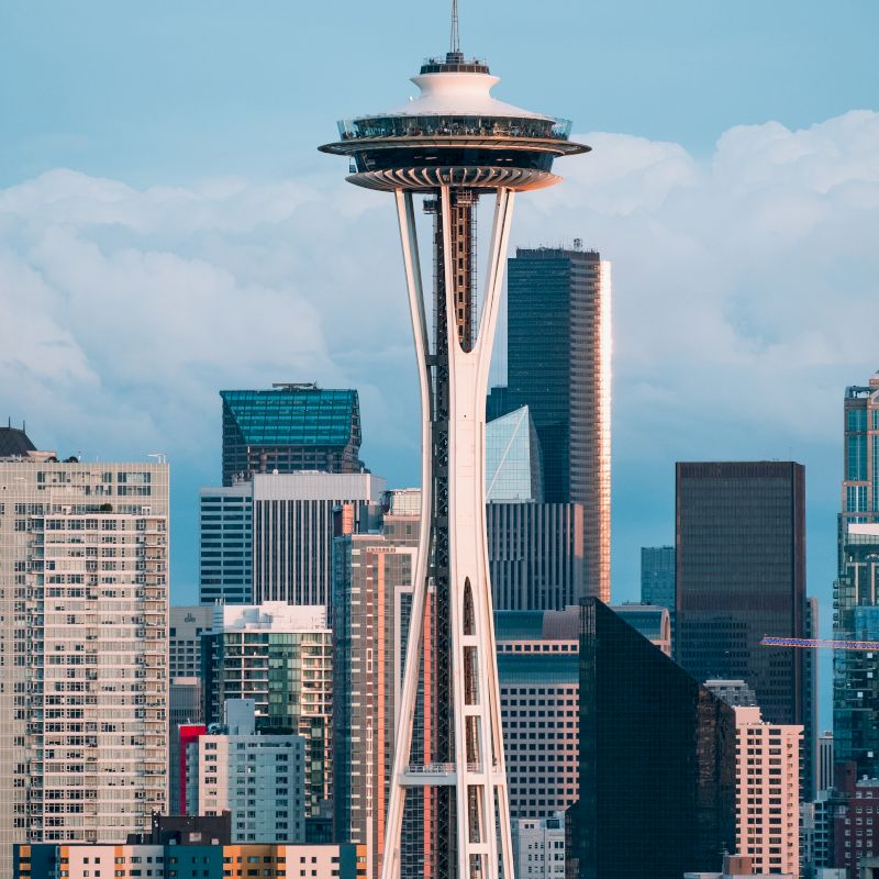 The image shows the Space Needle against a city skyline with various skyscrapers and a cloudy sky in the background.