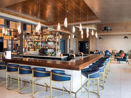 A modern, stylish bar with a central counter, blue barstools, hanging lights, and a well-stocked liquor shelf is shown.